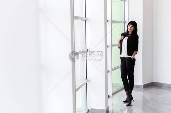 年轻有吸引力的快乐黑发女人穿着黑色西装站在办公室的窗户附近图片