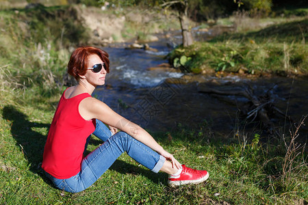 太阳镜中的年轻女子在阳光明媚的秋天坐在小溪图片