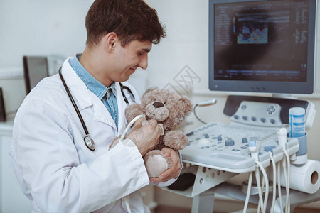 英俊开朗的男医生在毛绒玩具泰迪熊上使用超声波扫描仪图片