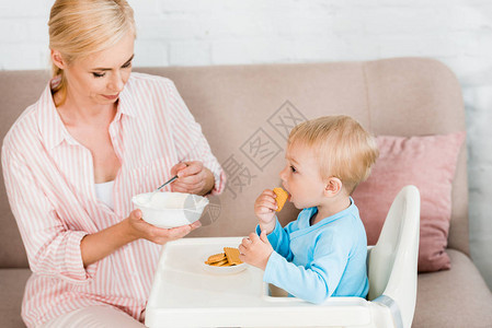 金发母亲拿着勺子和婴儿食物在可爱的小孩儿子旁边图片
