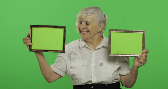 一位老妇人拿着两帧绿色图像一件白衬衫的老俏丽的祖母放置您的徽标或文本色度键绿屏背景图片