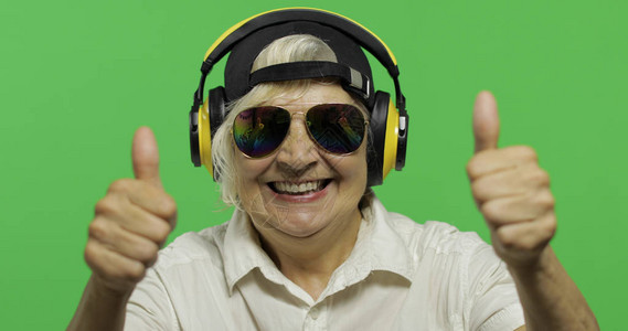 一位老妇人戴着耳机听音乐竖起大拇指太阳镜和盖帽的老相当愉快的祖母放置您的徽标或文本色度图片