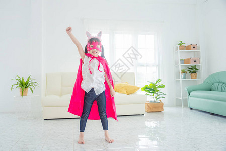 快乐的亚洲小女孩穿着粉红色的超级英雄服装站在客厅地板上准备回图片