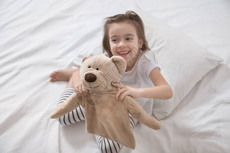 坐在白床上玩软熊玩具的可爱小女孩儿童睡眠和发育的概念背景图片
