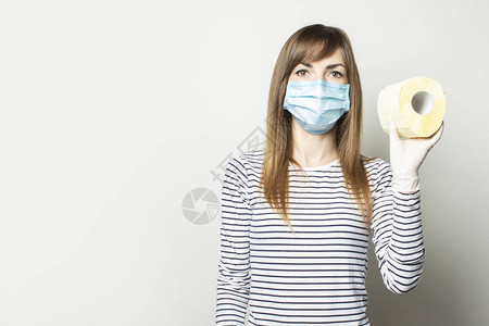 身戴医疗面具和医疗手套的年轻女孩在浅薄背景上持有一卷卫生纸图片