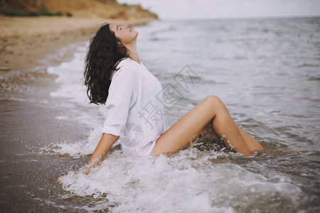 快乐的年轻女子穿着白衬衫坐在海滩上溅起浪花图片