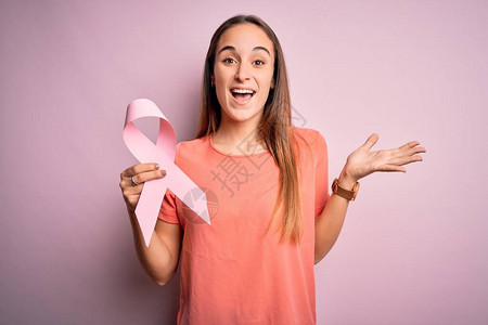 年轻漂亮的女人拿着粉红色的癌症丝带站在孤立的粉红色背景上图片