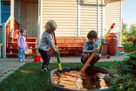 孩子们在乡间别墅背景的花园里玩耍一个女孩和一个男孩正在用儿童泵从一个小池塘里抽水三姐站在一图片