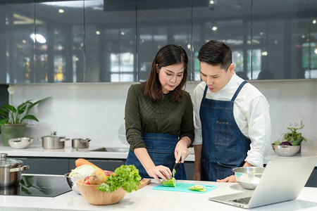 亚洲夫妇在家厨房一起做饭女切片蔬菜和笔记本电脑烹饪年轻夫妇正在图片