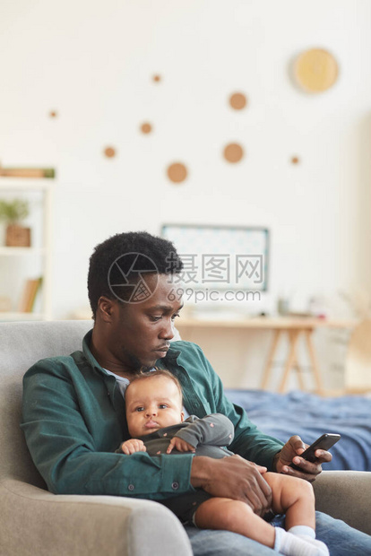 年轻非裔美国父亲坐在舒适的扶手椅上并使用智能手机复制空间时图片