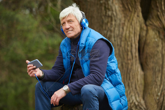 活跃老人在秋季森林徒步时坐在树旁休息时通过智能手机听音乐的肖图片