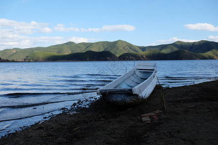 一条白色的小船搁浅在黑土海滩上蓝色的湖水在四周荡漾青山背景晴朗的蓝天在中背景图片