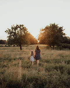 欧美母女欧洲家庭母女手牵着手沿着高的绿草走向夏天的日落草地和高大的树木背景