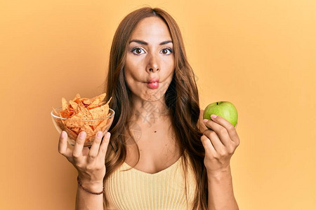 年轻的黑发女人拿着玉米片和健康的绿苹果用嘴斜眼疯狂和滑稽图片