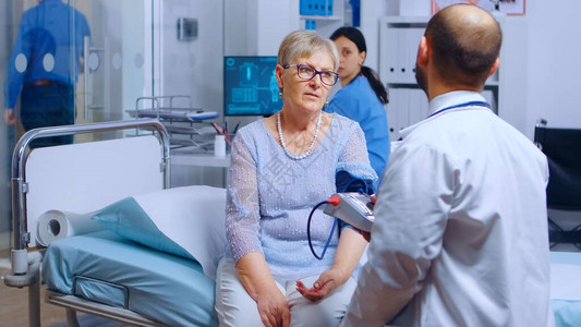 医生在后台工作的护士用监视器测量老年退休妇女的血压医疗保健医疗系统疾病检查背景图片