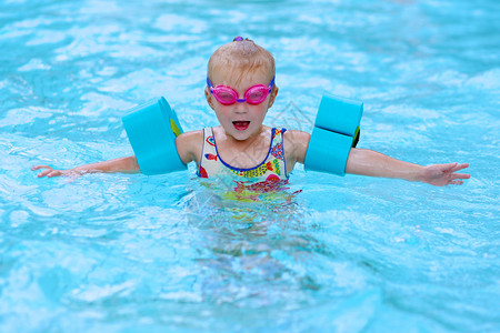 小孩享受游泳池可爱的蹒跚学步的女孩穿着五颜六色的泳衣粉红色的护目镜和蓝色的臂章在水中玩乐可爱的运动员孩子提倡健背景图片