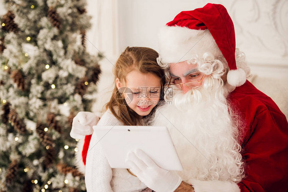圣诞节快乐女孩圣塔在数字触摸屏幕平板电脑烟囱和树彩色灯光上图片