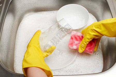 女手戴黄色防护橡胶手套用粉色清洁海绵对着装满泡沫和餐具的厨房水槽清洗图片
