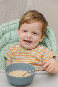 穿着条纹T恤的金发小男孩用勺子从灰色盘子里吃粥背景图片