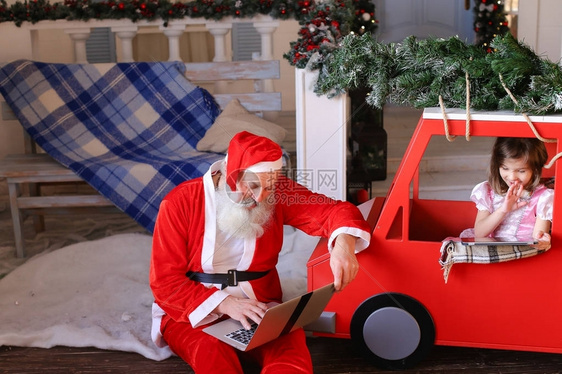 欢乐的圣诞老人在装饰的房子附近竖起大拇指快乐漂亮的女孩满意地在儿童车顶上买了绑的枞树圣诞装饰品图片