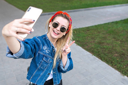 时尚快乐的女孩在太阳镜牛仔夹克和围巾站在公园采取自拍智能手机和微笑微笑的妇女摆在户外的相机智能手机公园背景图片