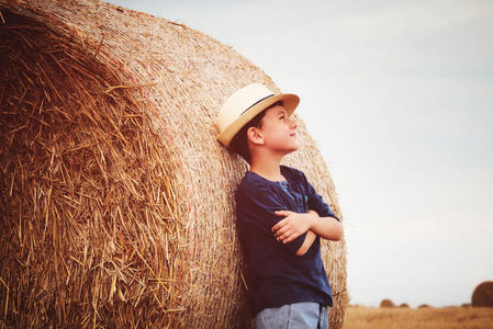 可爱的乡村男孩在附近草垛在日落时在夏天的帽子。夏天的概念，一个假期。与温暖的夏日儿童户外活动休闲