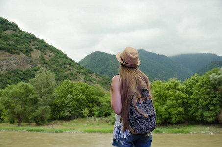 女孩与一个背包和一顶帽子，欣赏绿山川河流。查看从背面