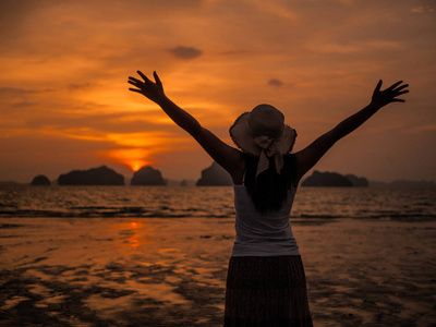 女人在海边戴着顶帽子下日出的张开双臂的轮廓。夏天的概念