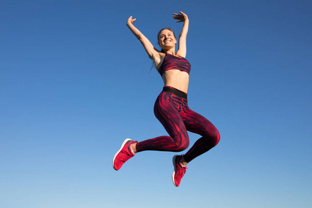 孤立的年轻快乐女运动员在运动服跳跃和飞行在蓝色天空背景。她享受夏天。健康的生活方式的概念，体育活动