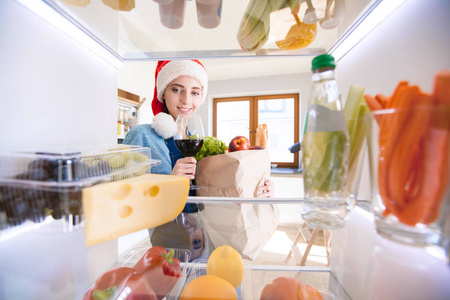 圣诞老人的帽子站附近打开冰箱充分的健康食品，蔬菜和水果的女性的肖像