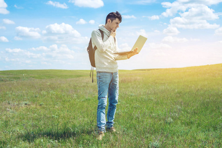 年轻人坐在一个绿色的草甸与笔记本电脑无线蓝多云的天空为背景