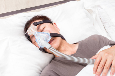 中年亚裔男子睡在他床上，穿着 Cpap 头饰 m