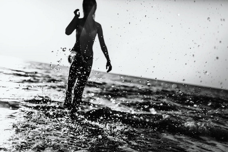 背光拍摄的在夏天阳光明媚的日子与她的双腿在海滩水中跑步的女孩。水溅