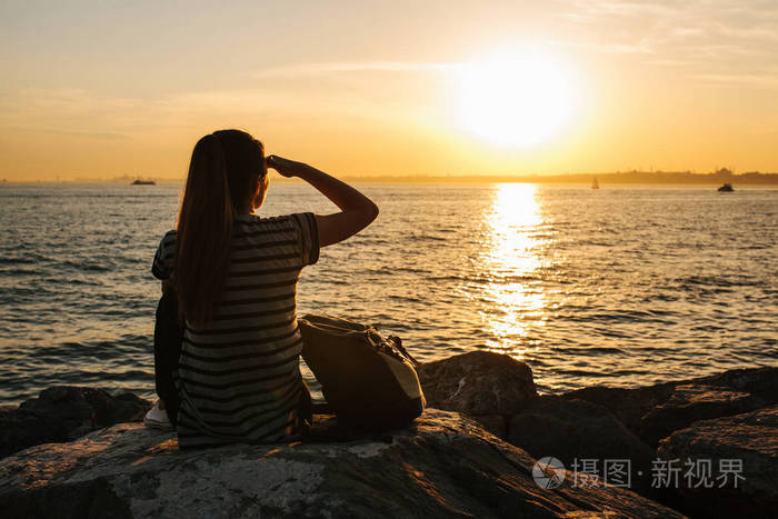 年轻游客的女孩背着日落时分坐在海边岩石上眺望远方休息放松旅行度假