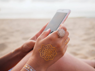 在海滩上的手里拿着智能手机的闪存纹身的女人。波西米亚风格。美丽的配件