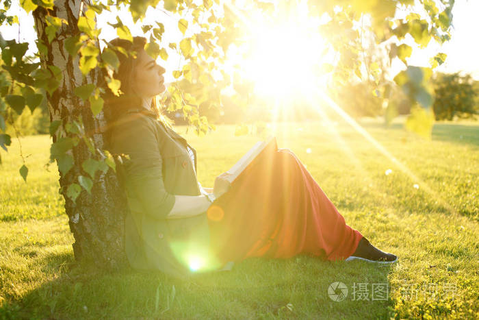 年轻女子拿着一本书在一棵树下坐在阳光灿烂的日子