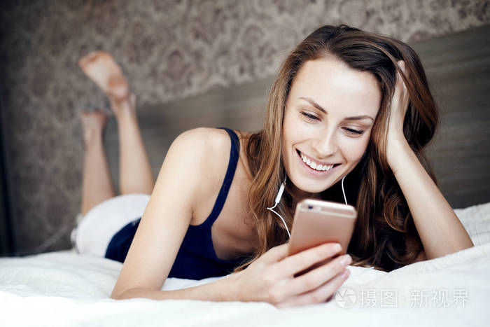 年轻女孩在卧室里听音乐的耳机和拿着手机在床上