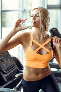 体育的金发碧眼女人正在喝水在健身房的跑步机