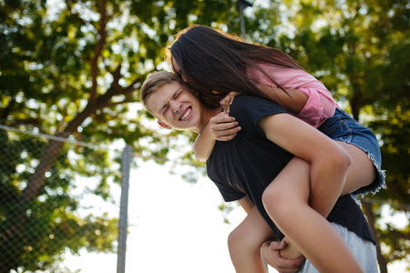年轻微笑的男孩抱着漂亮的女孩在他的背上和她玩的肖像, 而在公园花时间