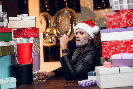 时髦的圣诞老人穿着黑色皮夹克抽雪茄, 喝威士忌。他坐在新年礼物周围