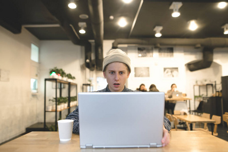 一个年轻人在咖啡馆工作, 在笔记本电脑屏幕上看起来很惊讶。学生在舒适的咖啡馆里使用互联网
