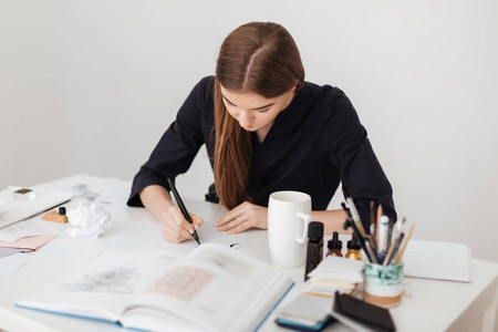 年轻美丽的女士坐在白色的办公桌上的肖像, 并在纸上写笔记与开放的书孤立