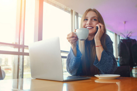 快乐的年轻时髦的女人与杯 cofe 交谈在电话里在咖啡馆。桌上的笔记本电脑