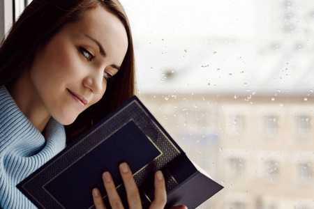 年轻的黑发女子穿着蓝色羊毛衫, 在雨中的房间里, 在窗台上读一本有趣的书, 多云和寒冷的天气