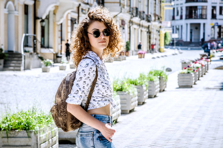 一个头发卷曲的女孩在黑色的圆形眼镜和棕色的背包在夏天的城市漫步