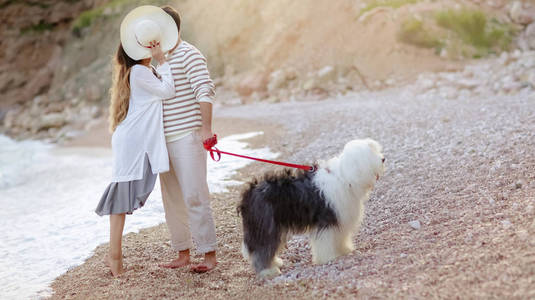 满长的年轻夫妇站在海滩上毛茸茸的狗和亲吻隐藏在帽子后面