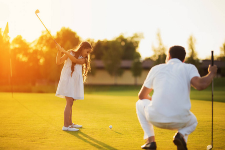 一个男人蹲在高尔夫俱乐部, 看着一个女孩谁是摆动俱乐部击球