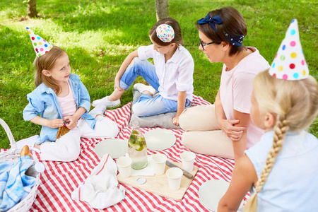女孩在生日帽和他们的母亲或老师谈话在公园的生日野餐