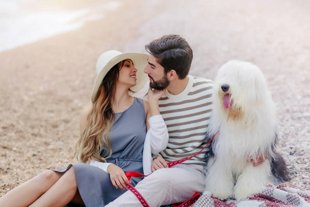 年轻夫妇在爱休息与毛茸茸的狗坐在沙滩上的格子上, 微笑着看着对方