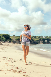 在日落时在热带海滩上运行的太阳镜的运动和健康的年轻女子。印度尼西亚巴厘岛